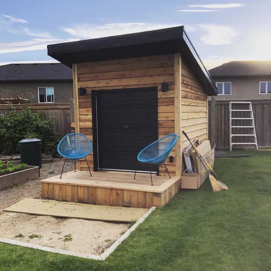 Small wooden shed with a black door, two blue chairs on the porch, and a broom leaning against the side, surrounded by a garden