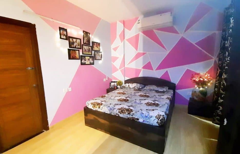 Bedroom with geometric pink and white accent wall, double bed with floral bedding, family photos, fairy lights, and air conditioner