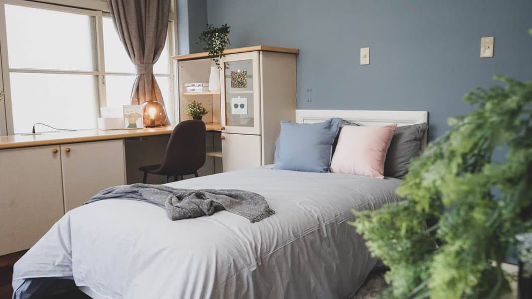 Bright bedroom with a neatly made bed, blue and pink pillows, a desk with a chair, shelves, and a window with curtains