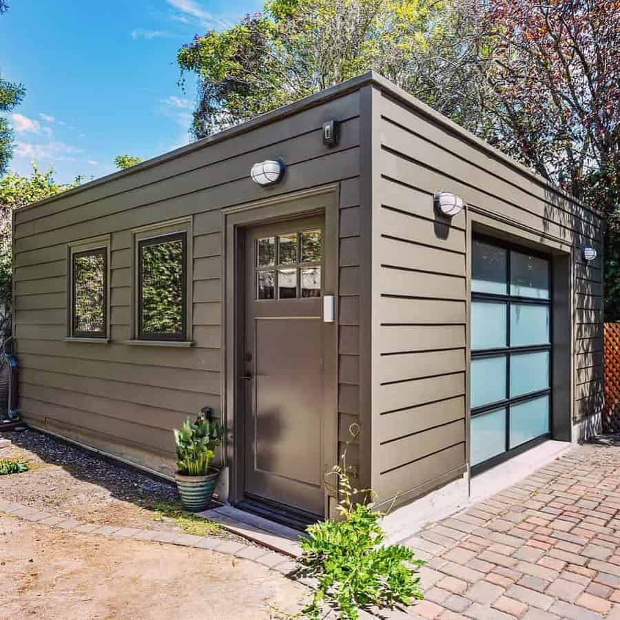 Modern shed with an olive-green entry door, frosted glass garage door, and matching siding, surrounded by greenery and brick paving