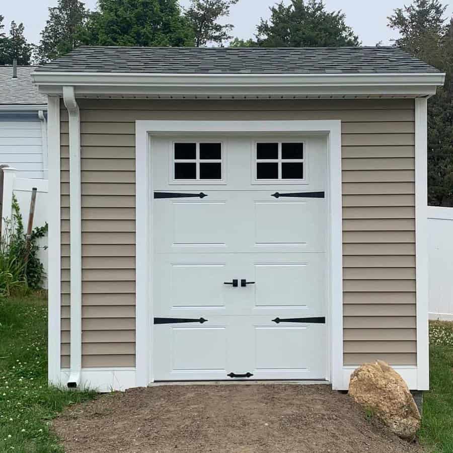 Small beige shed with a gray shingled roof, white trim, and a white door with windows, rock in front on the gravel path