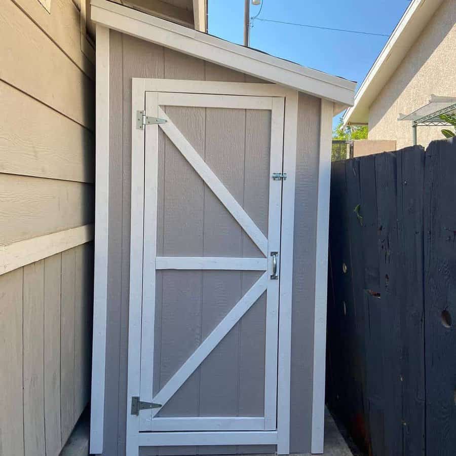 Narrow wooden storage shed with a slanted roof and a white-framed door, situated in a tight space between two buildings