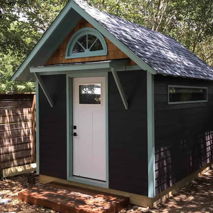 Small wooden shed with a pointed roof, white door, and teal trim, surrounded by trees