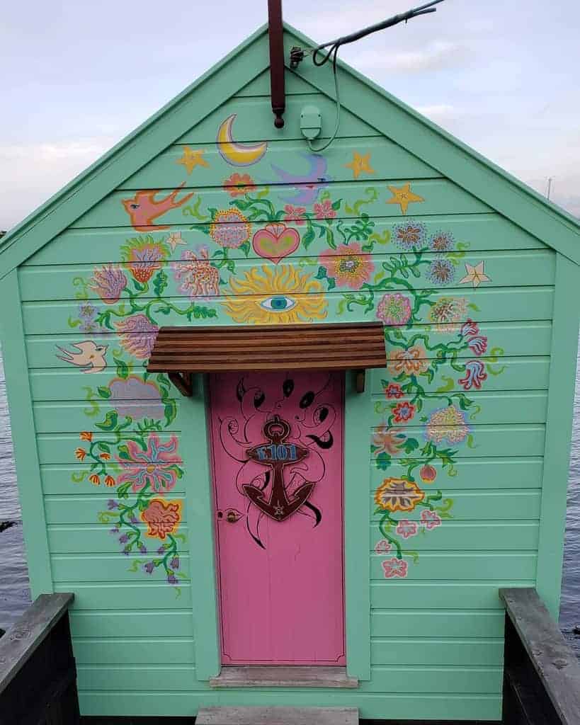 A small green shed with a pink door, covered in colorful floral and nautical murals near a body of water