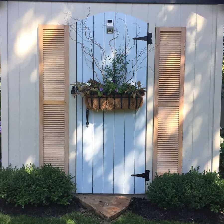 Light blue garden shed door with wooden shutters and a hanging basket of plants, surrounded by green shrubs