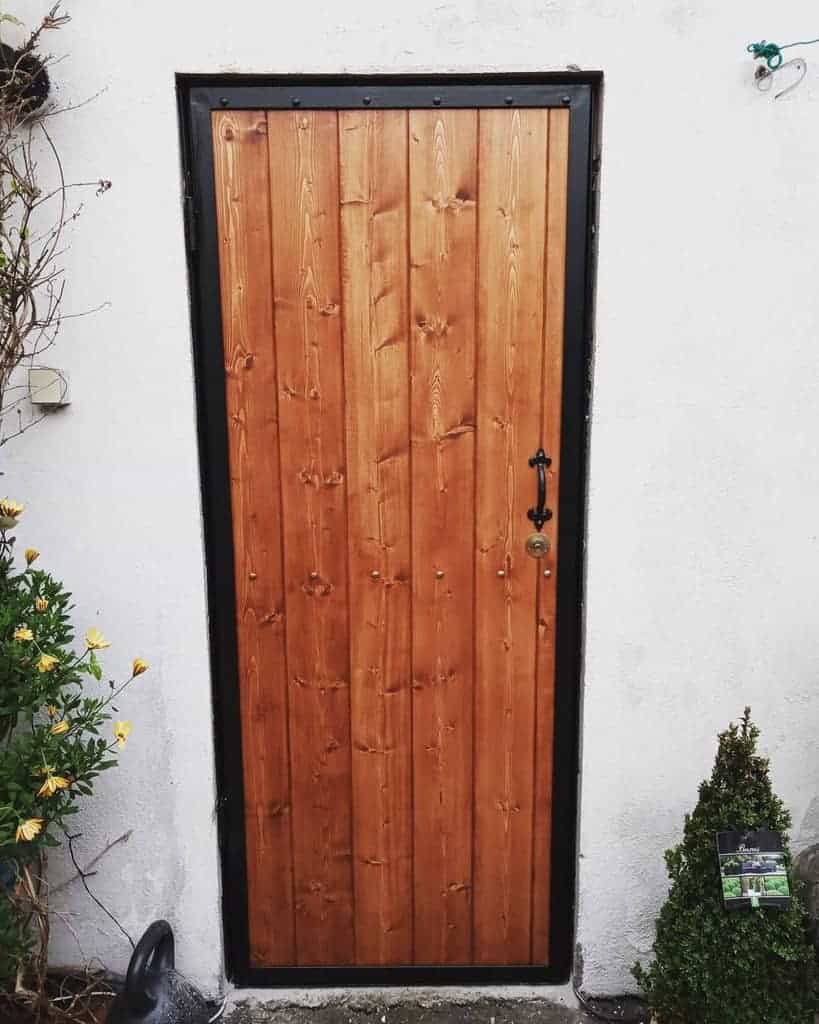 A wooden door with a metal frame set in a white wall, surrounded by small plants and a watering can on the left