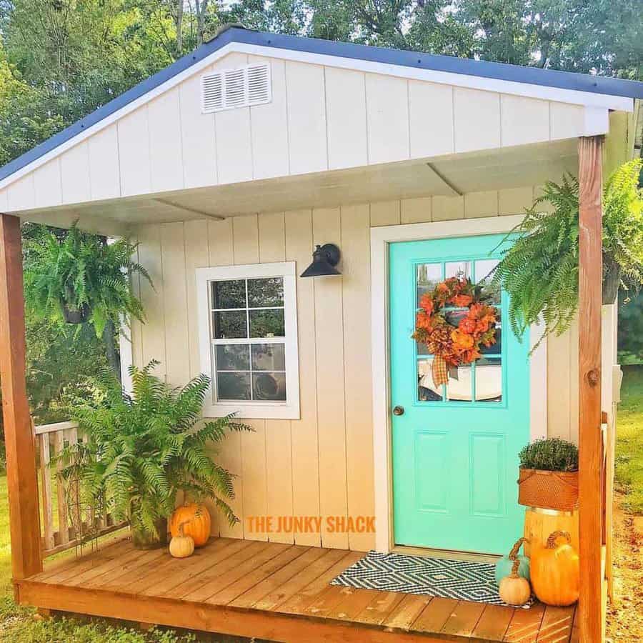 Charming shed with a bright turquoise door, fall wreath, and pumpkins on a wooden porch, surrounded by lush greenery and ferns