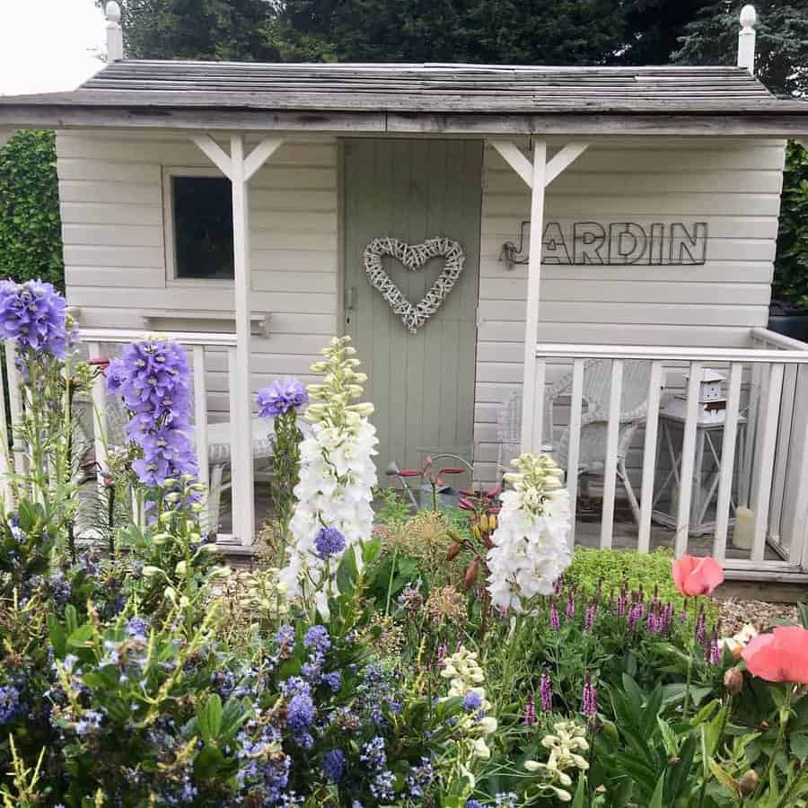 Small garden shed with white walls, a heart wreath on the door, and "JARDIN" sign surrounded by colorful flowers and greenery