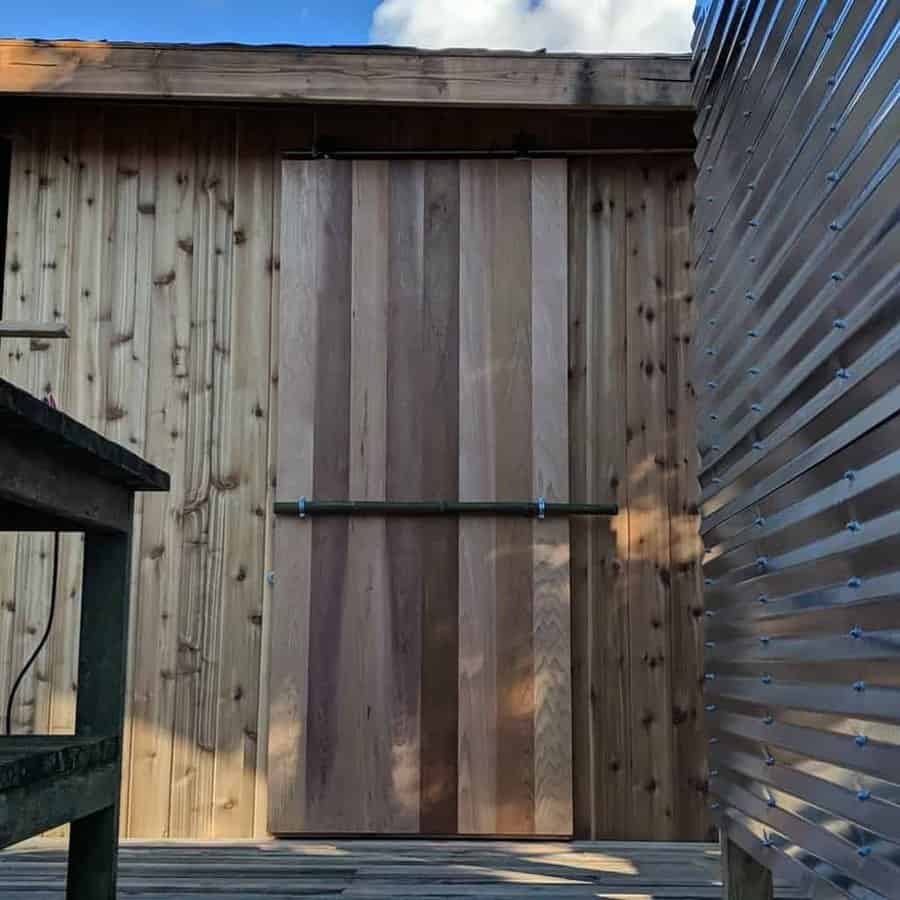 Rustic wooden shed with a vertical plank sliding door, metal track hardware, and a natural wood finish, set against a metal wall