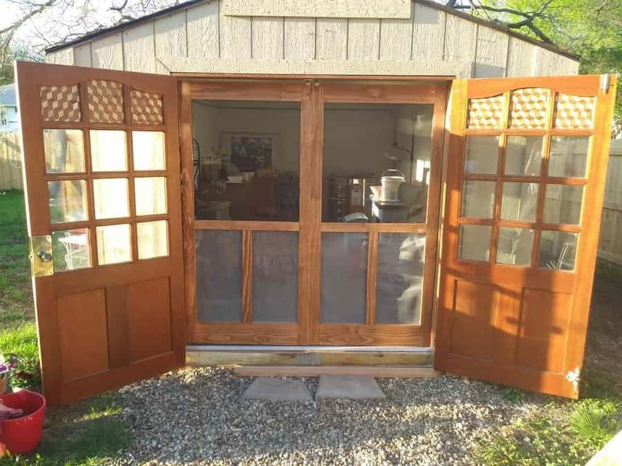 Wooden double doors open to a sunlit room inside a garden shed, with potted plants on the gravel outside