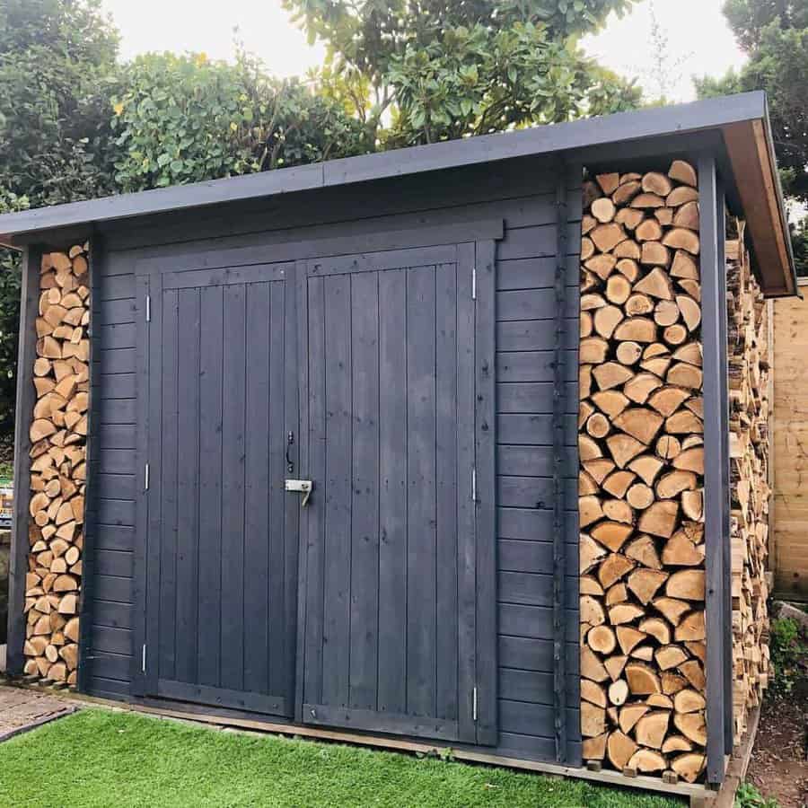 Dark wooden shed with double doors, secured with a latch, and built-in firewood storage on both sides, set in a green garden