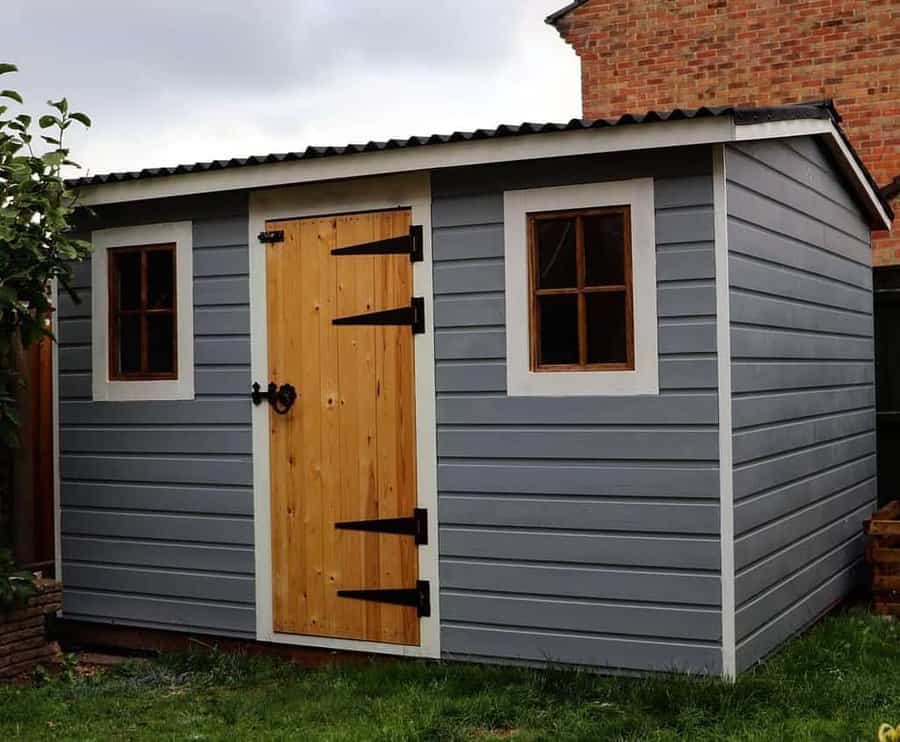 Gray shed with a natural wood door, black wrought iron hinges, and two small windows framed in white, set in a grassy backyard