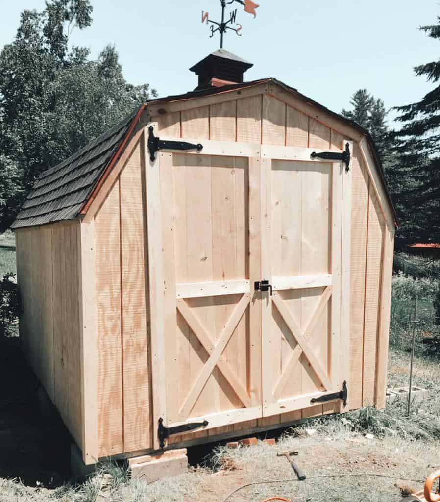 A newly built wooden shed with double doors and a weather vane on top, surrounded by trees and grass