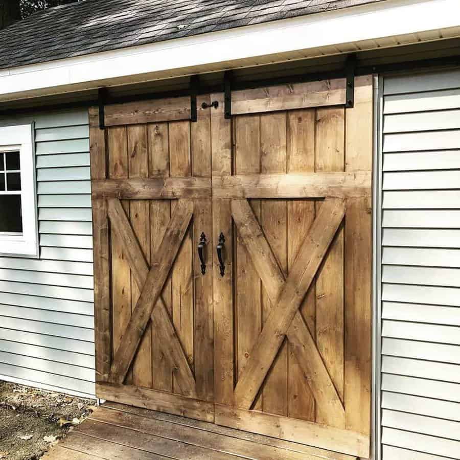 Rustic wooden barn-style sliding doors with black metal hardware on a light green shed with white trim, set on a wooden deck