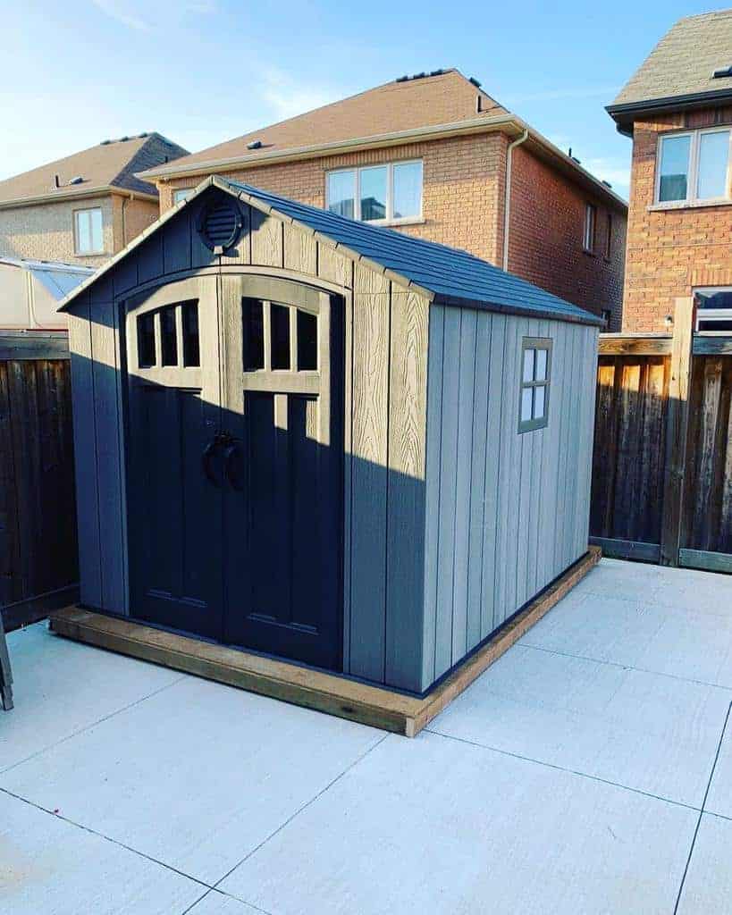 Modern gray shed with black double doors featuring arched windows, set on a raised wooden platform in a fenced backyard