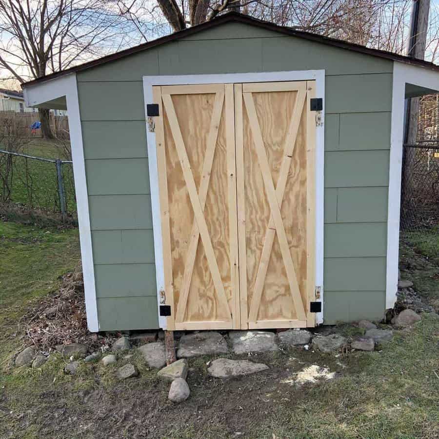 A small green shed with wooden double doors and a stone border at the base, set in a grassy yard with trees and a fence in the background