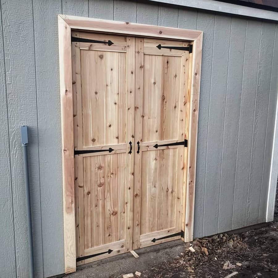 Natural wood double doors with black wrought iron hinges and handles, framed with matching trim on a gray shed exterior