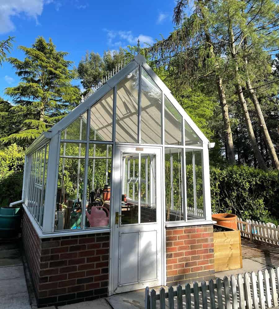 Glass greenhouse with white frame and brick base in a sunny garden