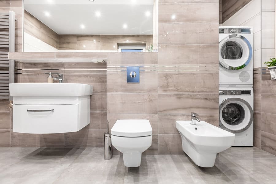 Modern bathroom with sink, toilet, bidet, and stacked washer/dryer against beige tiled walls and reflective mirror