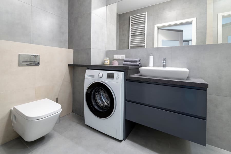 Modern bathroom with a washing machine, wall-mounted toilet, large mirror, sink, and grey cabinets