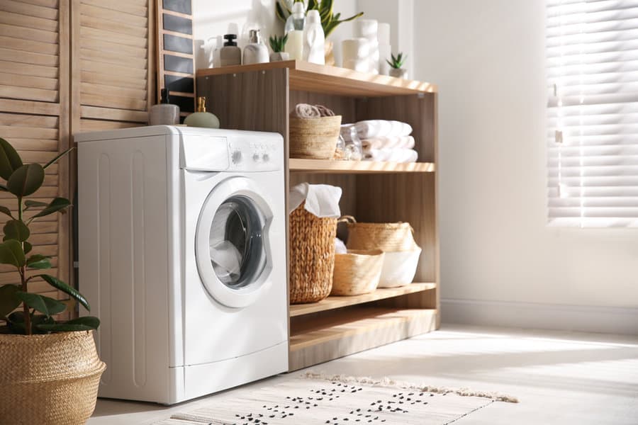 Modern laundry room with a white washing machine, wicker baskets, towels, and plants on wooden shelves by a sunny window