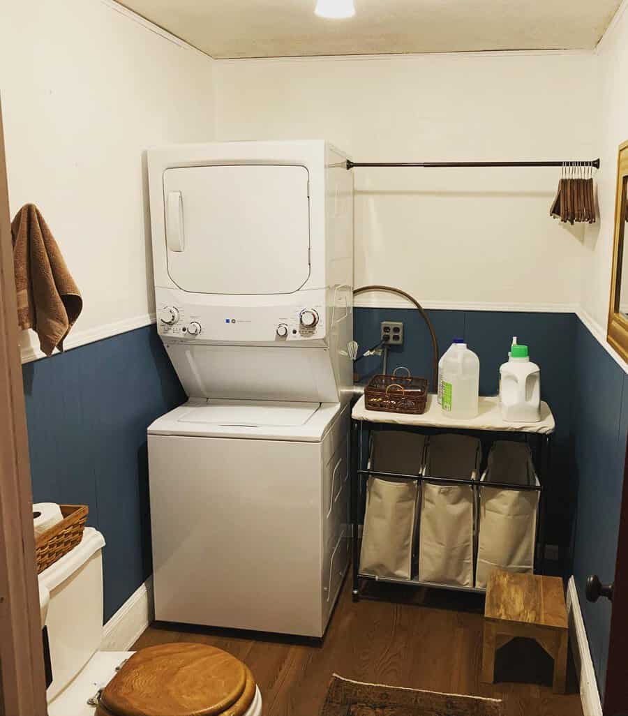 Small laundry room with stacked washer and dryer, a cart with laundry supplies, a drying rod, and a stool against white and blue walls