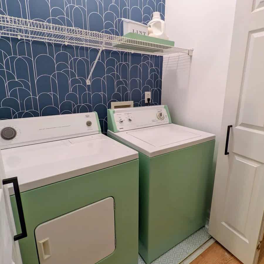 Laundry room with teal washer and dryer, blue patterned wallpaper, white shelf with items, and a partially open door