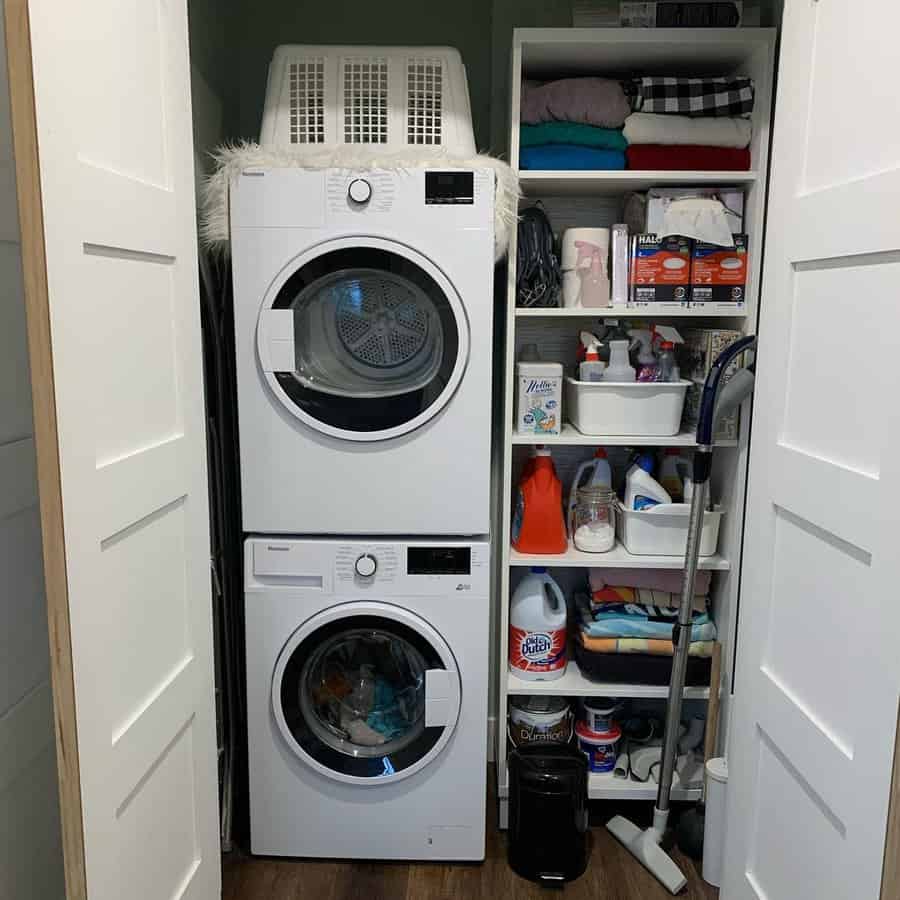 Compact laundry room with stacked washer and dryer, shelves of folded clothes, cleaning supplies, and laundry basket on top