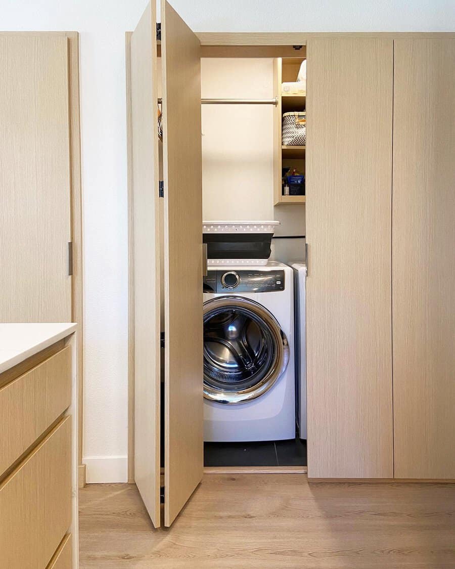 A front-loading washing machine is partially enclosed within a light wooden cabinet next to shelves with laundry items