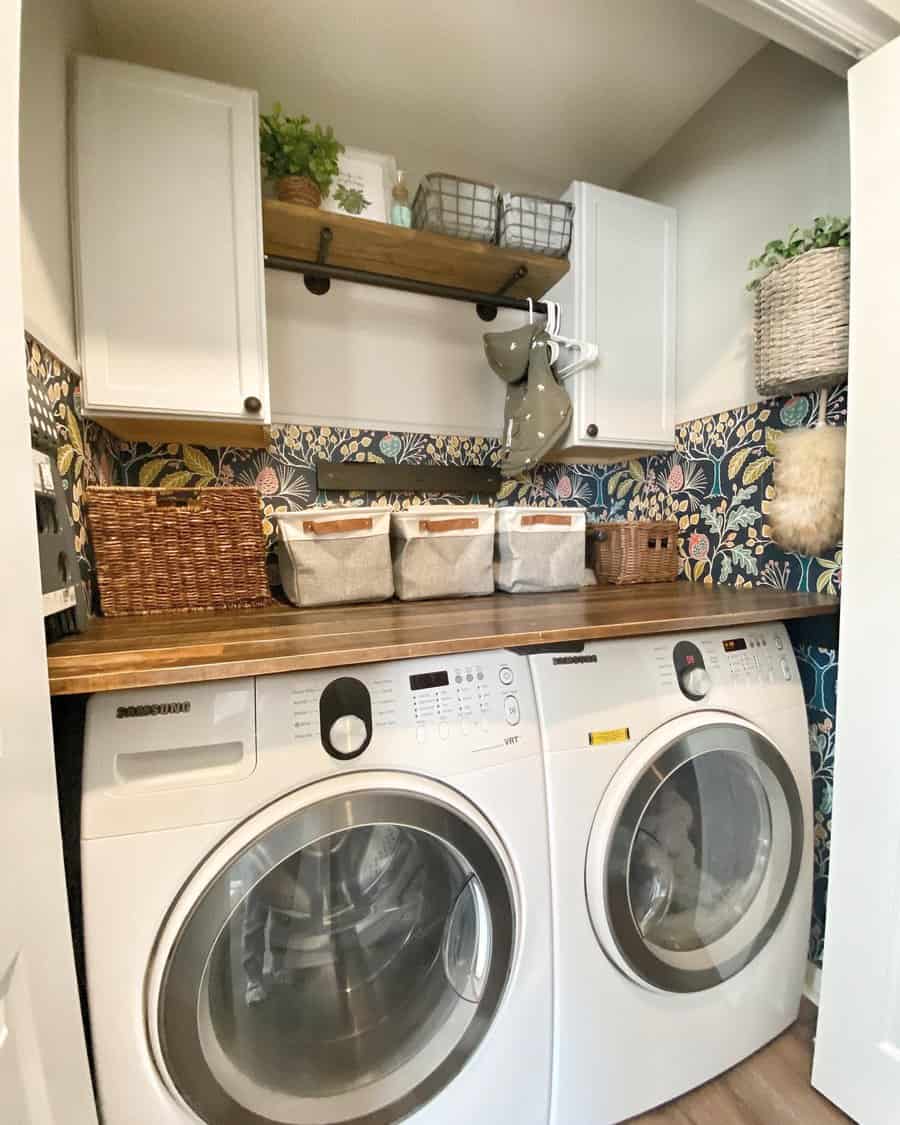 A compact laundry room with a washing machine, dryer, wooden countertop, baskets, and shelves with decorative items