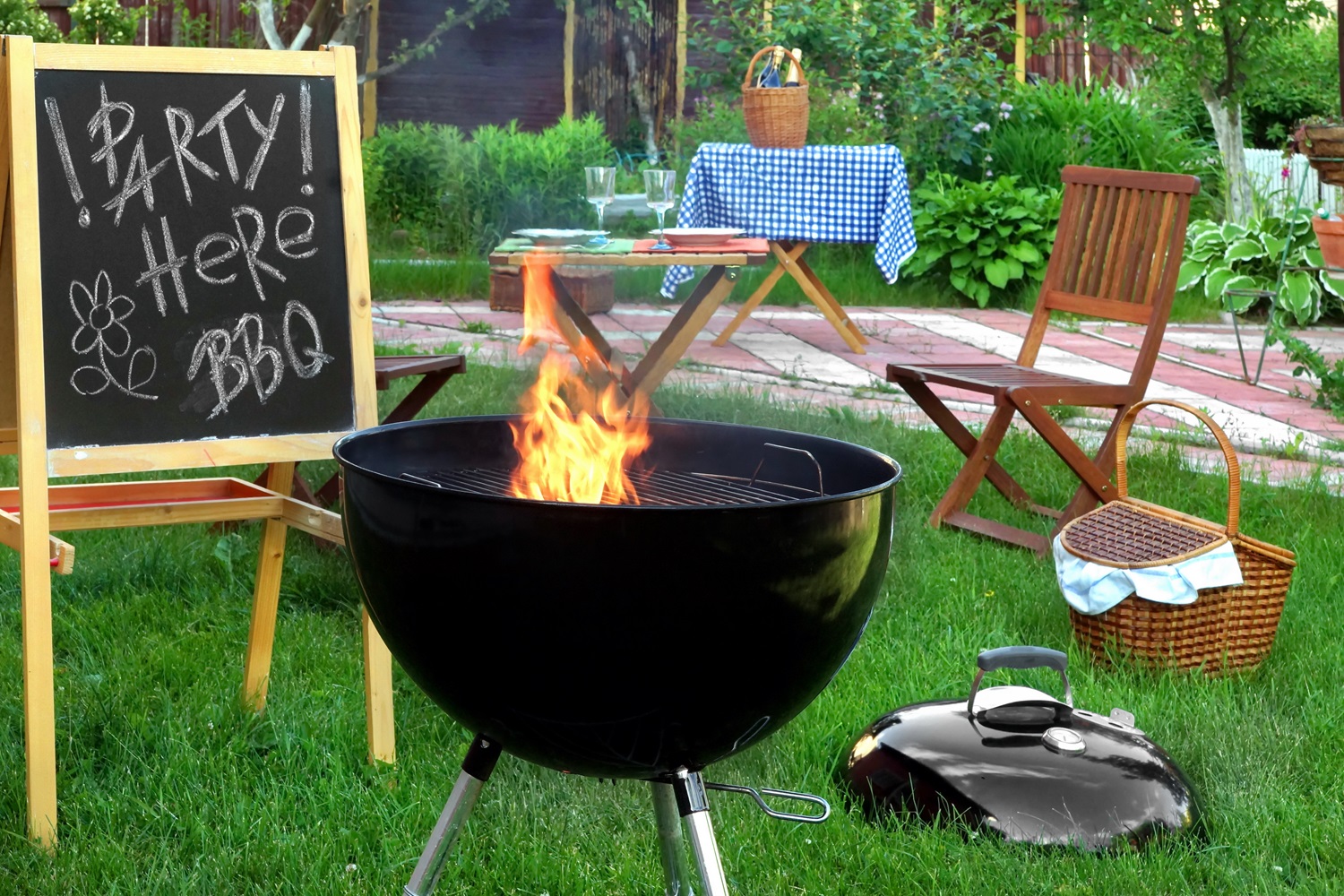 A backyard with a BBQ grill, sign reading "Party Here," table, chairs, and picnic basket.