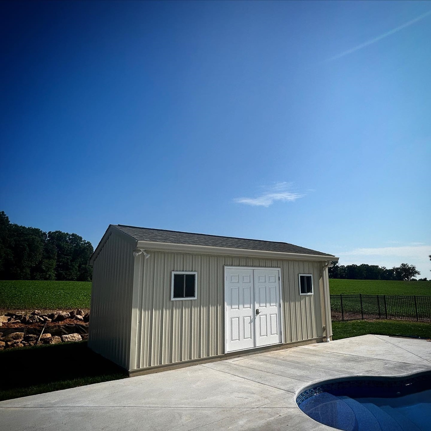 A small backyard white shed with double doors next to a pool under a clear blue sky.