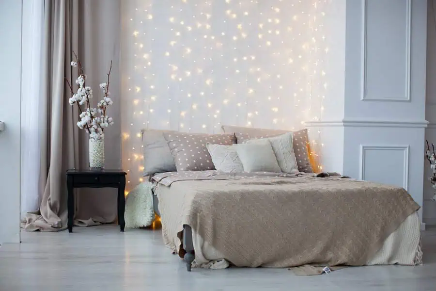 Cozy bedroom with fairy lights backdrop, beige bedding, and floral decor on a black nightstand.