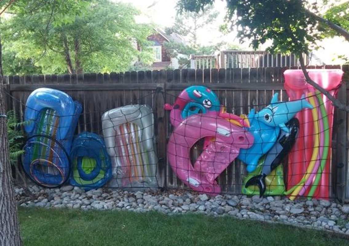 Inflatable pool toys, including floats, rings, and animal shapes, are neatly stored in a net against a backyard fence.