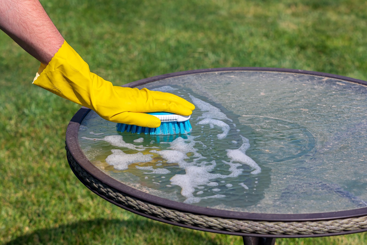 A hand in a yellow rubber glove scrubs a round glass outdoor table with a brush, creating soapy water. The table is in a grassy yard.