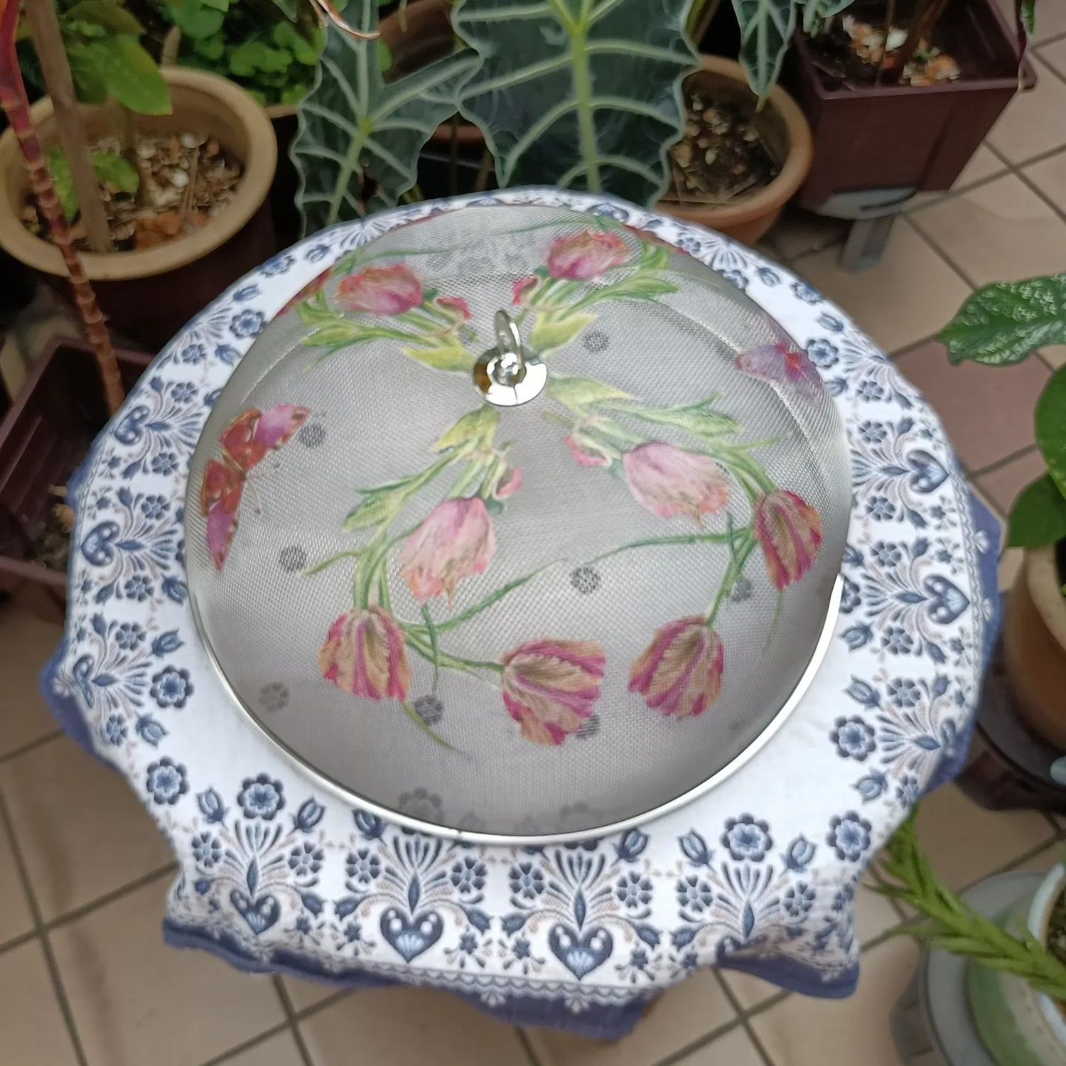 A mesh food cover with a floral design sits atop a blue and white tablecloth with heart-shaped patterns, surrounded by potted plants on a tiled floor.