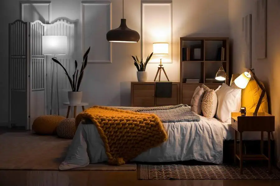 Cozy bedroom with warm lighting, wooden furniture, mustard yellow knit blanket, and various light fixtures.