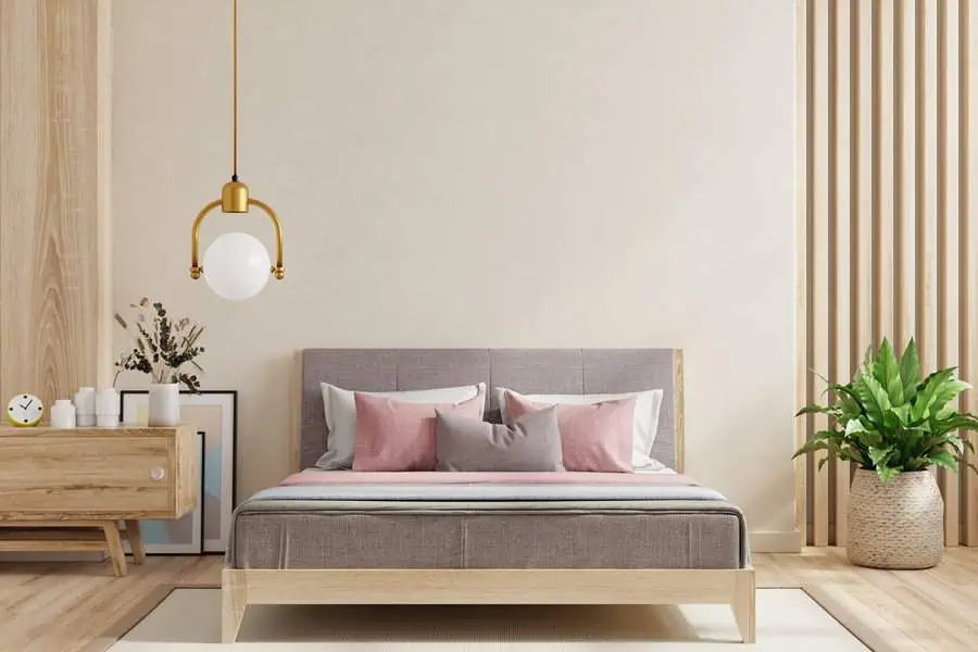 Minimalist bedroom with wooden furniture, grey upholstered bed, pink and grey pillows, modern pendant light, and a potted plant.