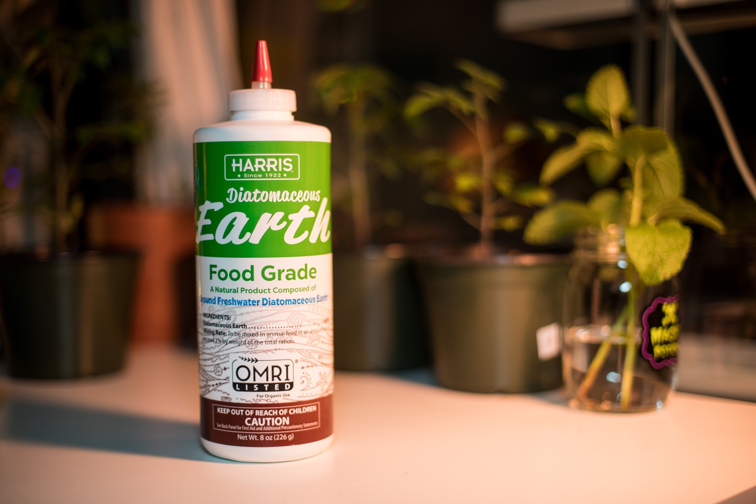 A bottle of Harris Diatomaceous Earth Food Grade placed on a white surface with potted plants in the blurred background.