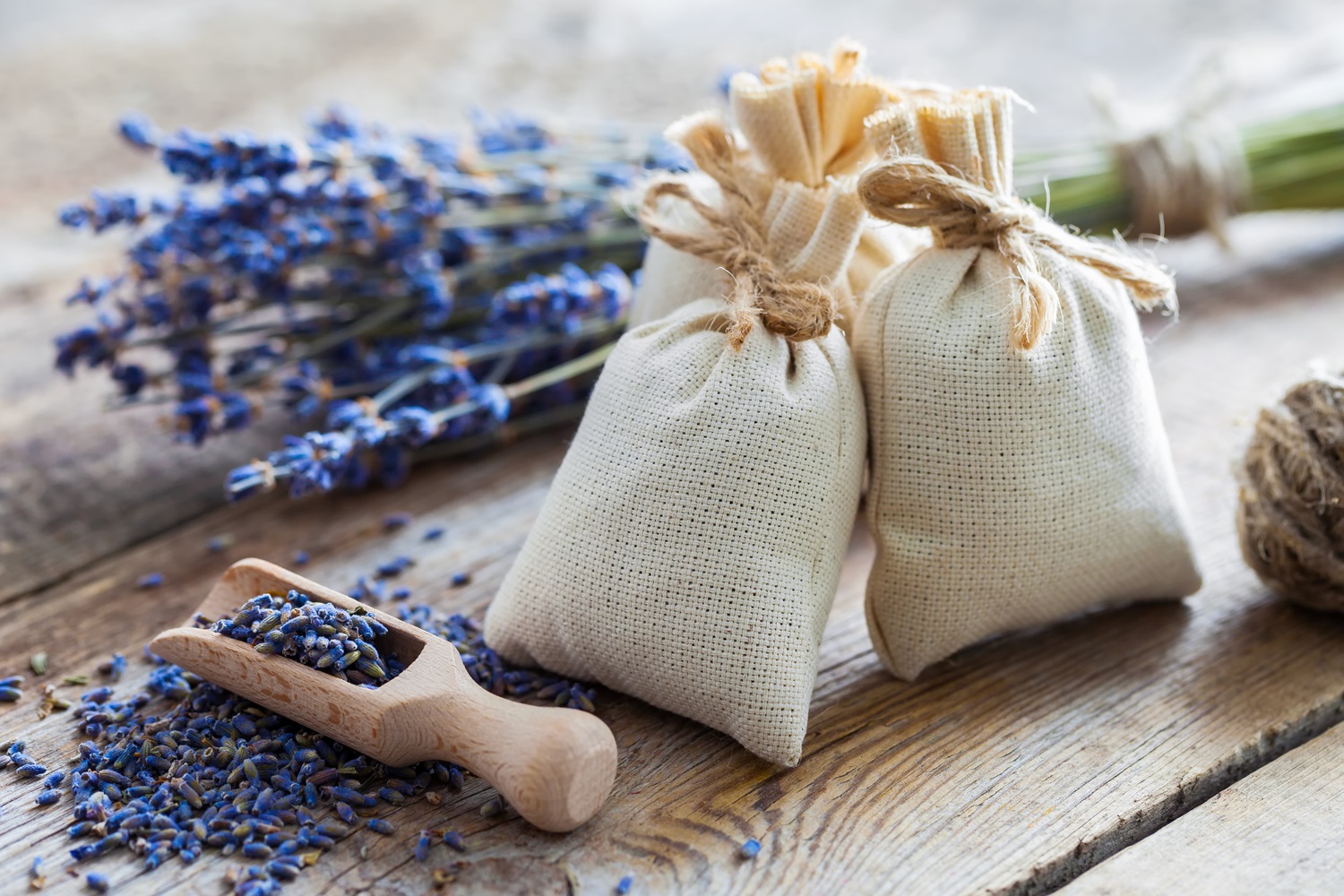 Three small burlap bags tied with string sit on a wooden table, surrounded by dried lavender sprigs.