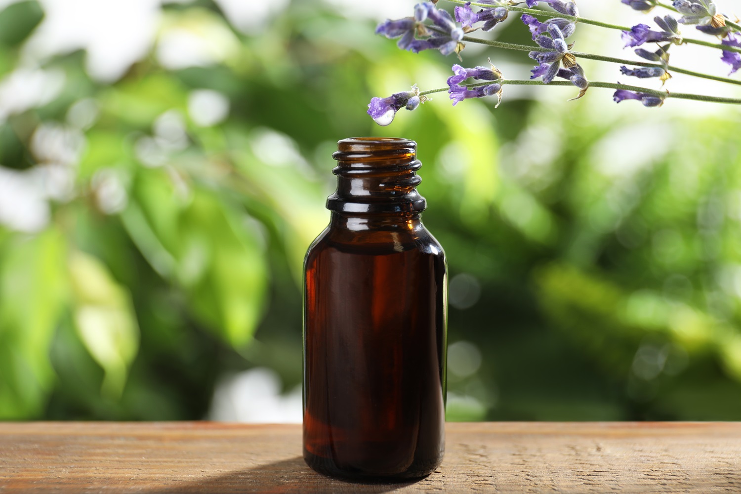 A small, brown glass bottle with an open top sits on a wooden surface. Lavender sprigs with purple blossoms hang above, and a vibrant green leafy background is blurred behind.