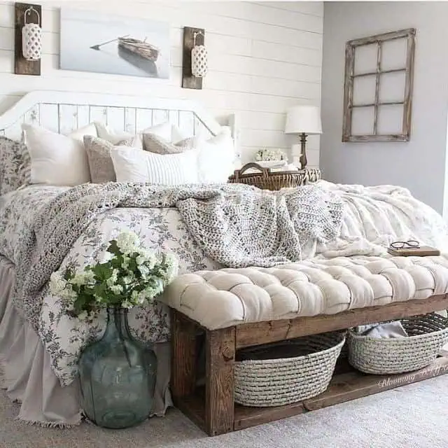 Cozy bedroom with a white shiplap wall, rustic wooden bed frame, tufted bench with storage baskets, and floral bedding.