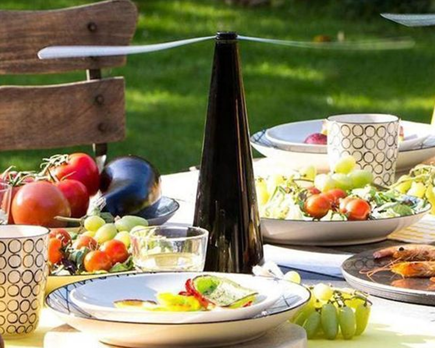 A sunlit outdoor dining table set with fly fan, salads, fresh tomatoes, grapes, an eggplant, cups, and a tall black pepper grinder.