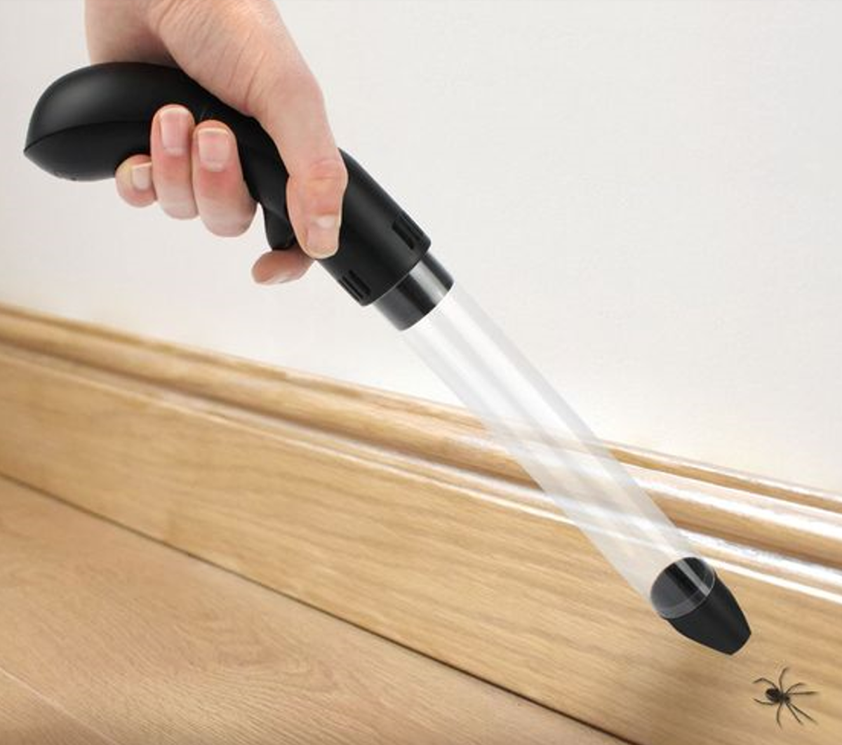 Hand holding a black and clear vacuum device directed at a small spider on a wooden floor.