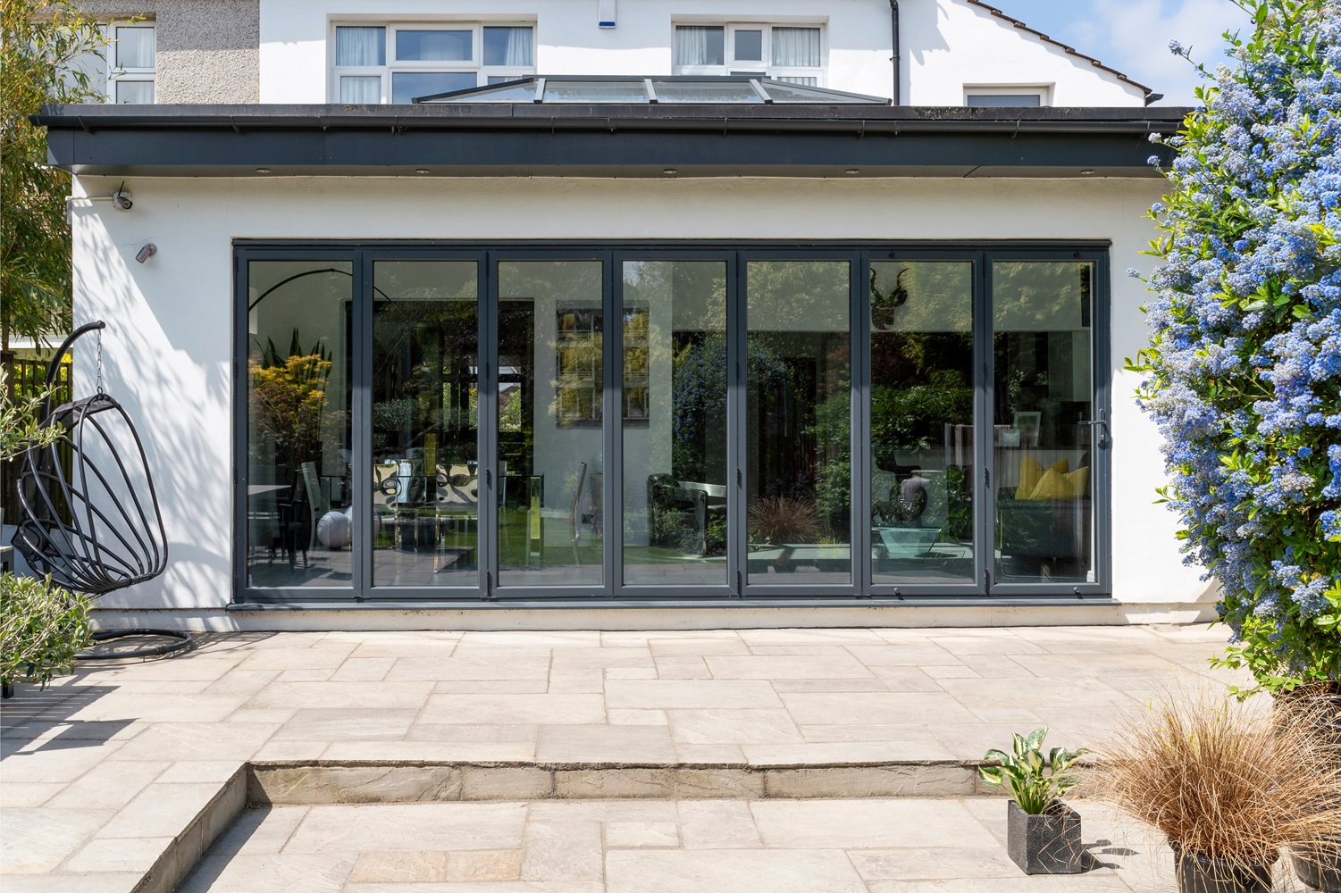 Modern house exterior with large, black-framed glass doors opening to a patio with beige tiles. 