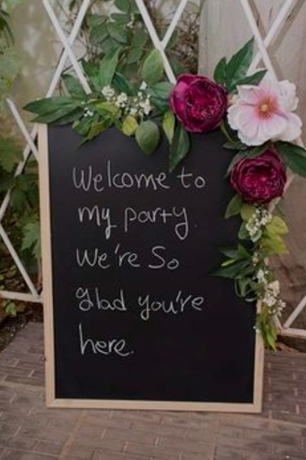 Chalkboard sign with flowers saying, "Welcome to my party. We're so glad you're here.