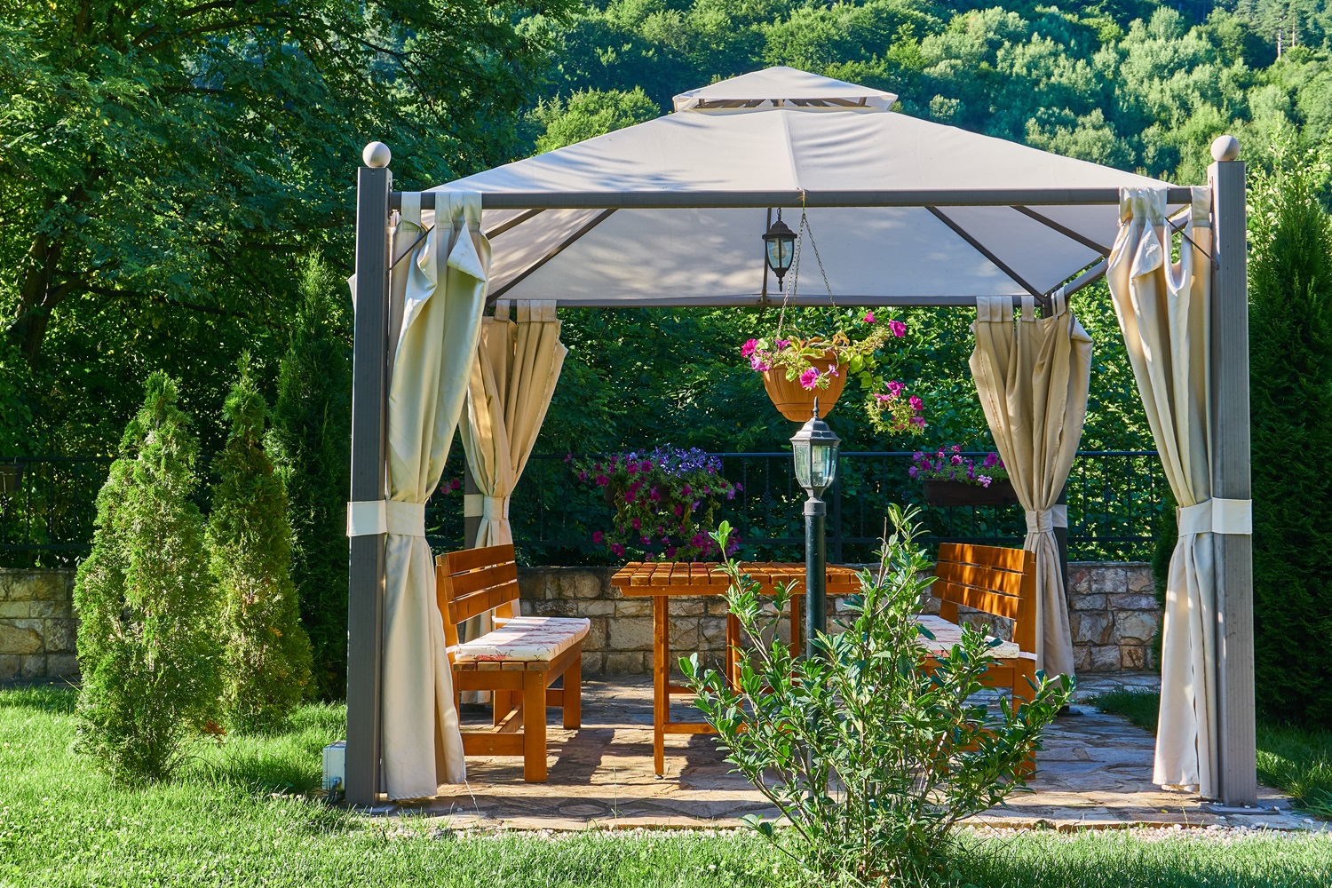 A gazebo with curtains in a lush garden, featuring wooden benches and hanging flowers