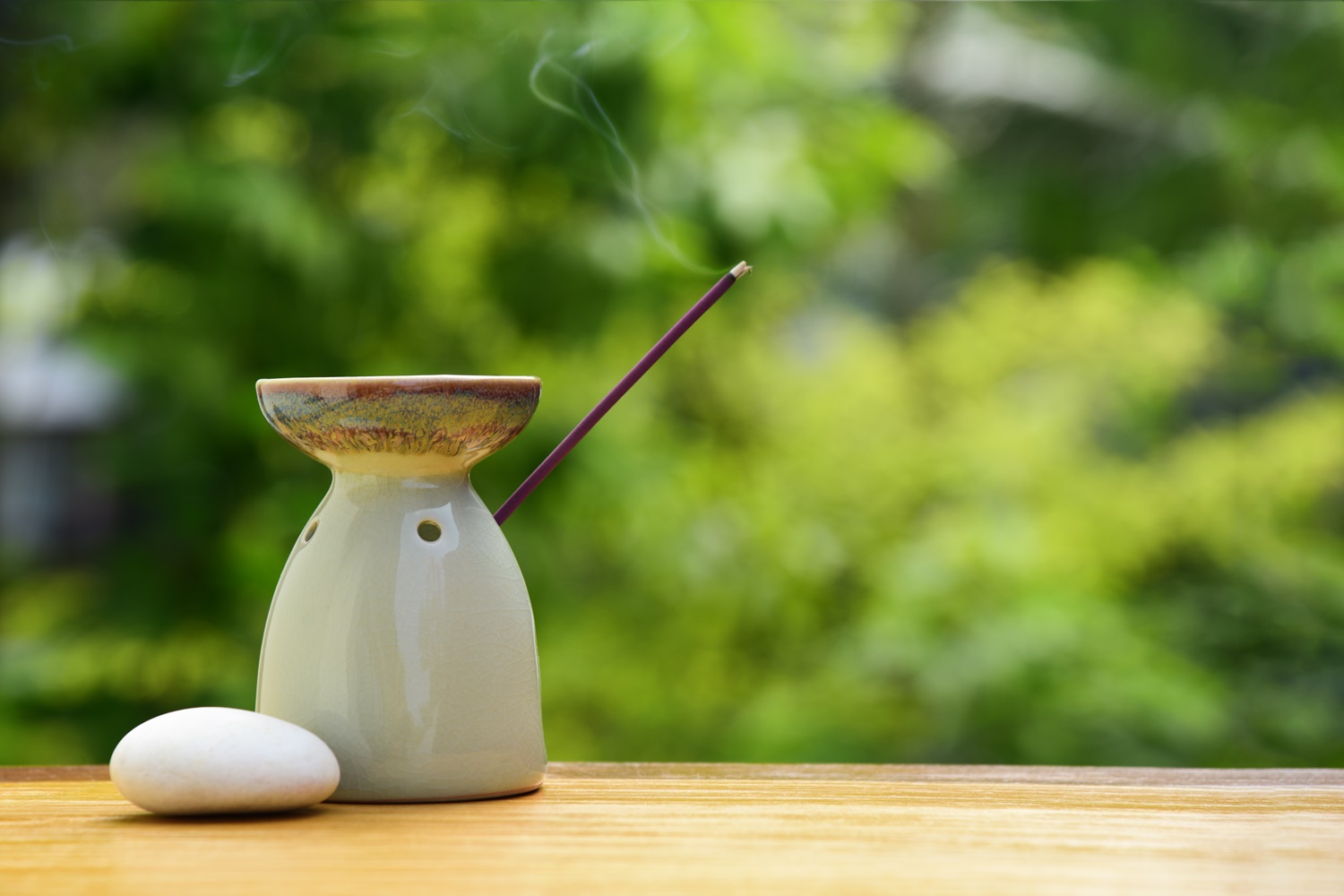 A white ceramic incense holder with a smoldering incense stick and a smooth white stone sits on a wooden surface against a green, blurred natural background.