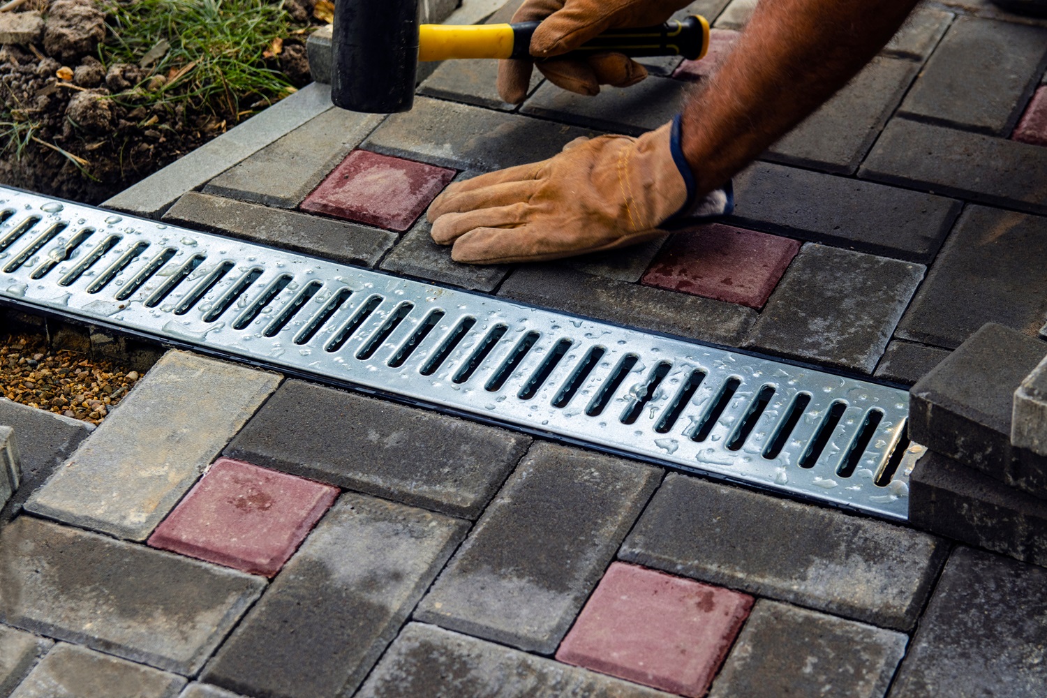 Person wearing gloves uses a rubber mallet to install interlocking concrete pavers around a metal drainage grate on a patio.