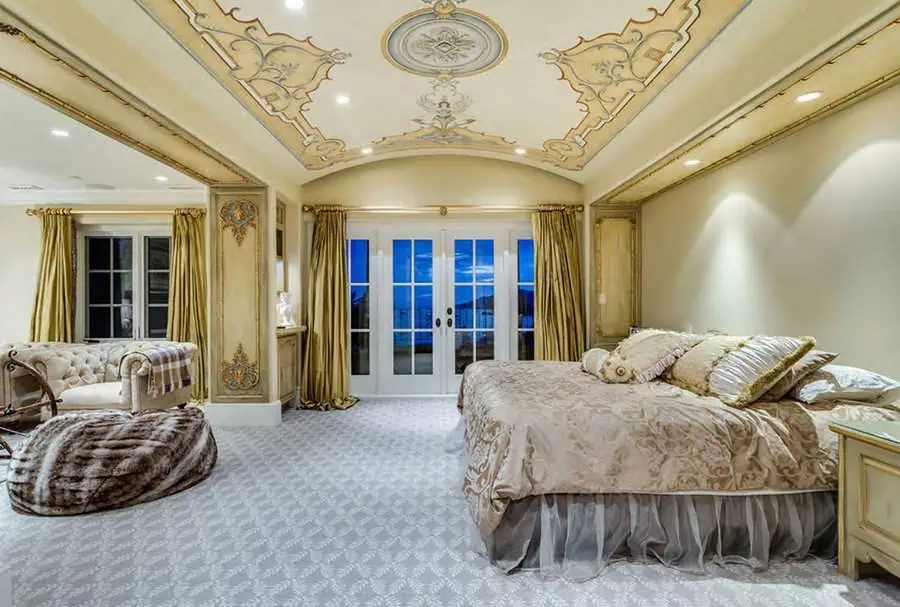 Luxurious bedroom with ornate ceiling design, elegant decor, French doors, and a plush chaise lounge.